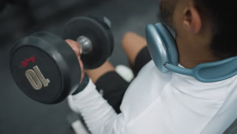 close up of gym coach wearing white top and blue headphones around neck while lifting dumbbell during intense arm workout session in modern gym