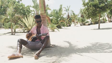 happy african american man sitting under tree playing guitar and singing on sunny beach, slow motion