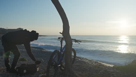 The-man-wears-his-gloves-and-backpack,-retrieves-his-mountain-bike-propped-against-a-tree,-and-sets-off,-leaving-behind-the-breathtaking-coastal-view-illuminated-by-the-morning-sun