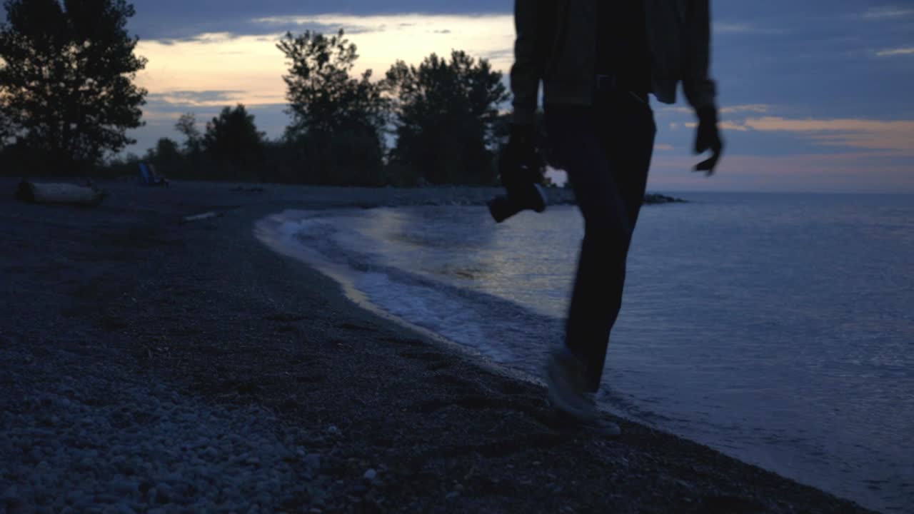 Man With Camera Walking By The Shoreline In The Early Evening - medium shot