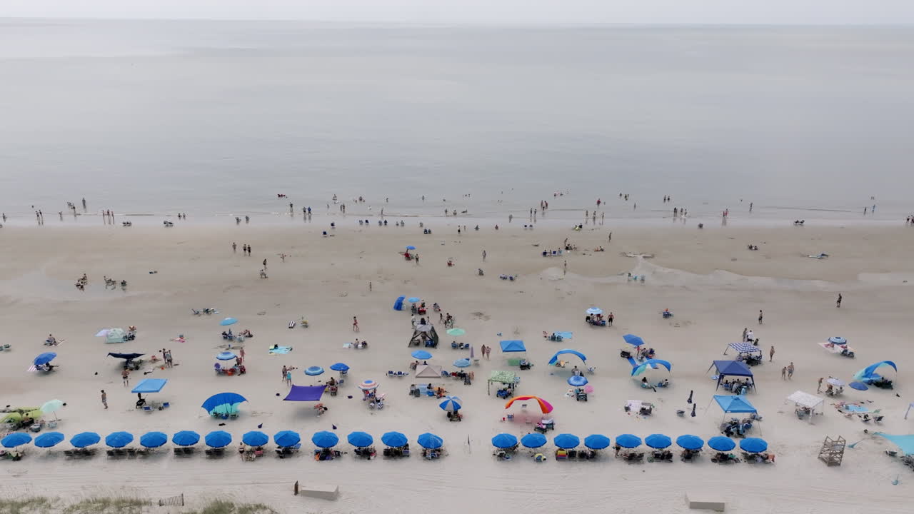 Beachgoers dot the sand and wade in shallow waters beneath a hazy sky, framed by rows of blue umbrellas along the coast