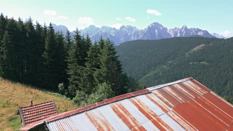 Drone-video-slowly-rising-vertically-from-behind-old-farm-barn-in-historical-hillside-town-to-reveal-forest-and-mountain-range-in-the-background-on-a-sunny-day-in-summer-in-the-Dolomites-Italy