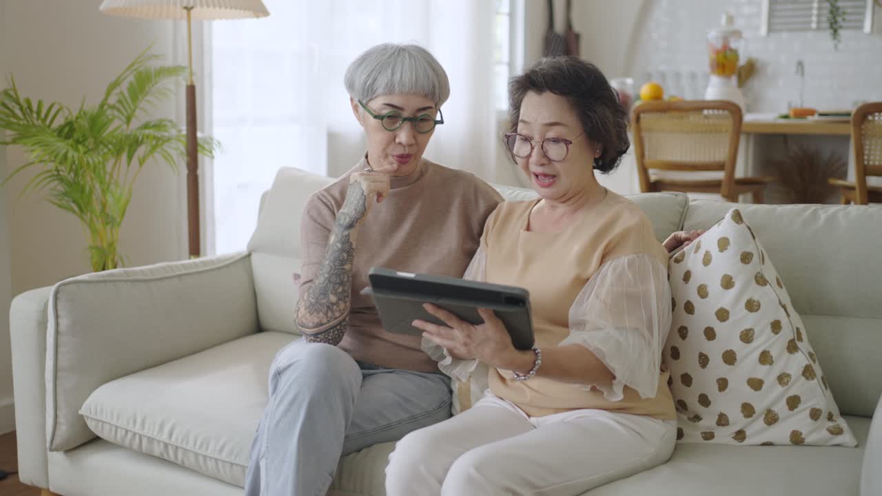 Due amiche anziane asiatiche in pensione sedute sul divano che usano un tablet insieme a casa.