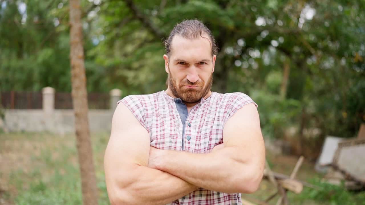 A serious man with a beard and plaid shirt standing outdoors with arms crossed