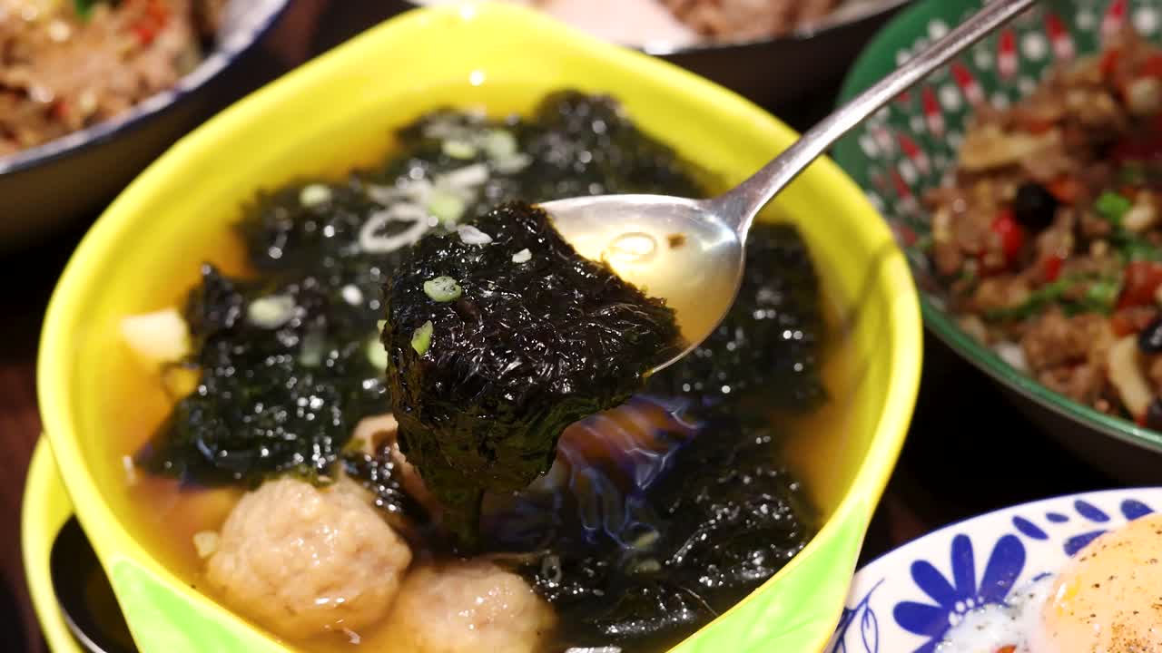 A spoon lifts seaweed from a yellow bowl of hot soup with pork meatballs, surrounded by vibrant side dishes under warm indoor lighting, close-up perspective