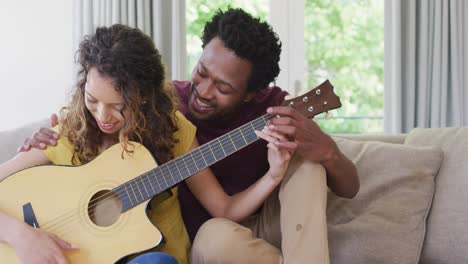 feliz pareja biracial sentada en el sofá juntos y tocando la guitarra