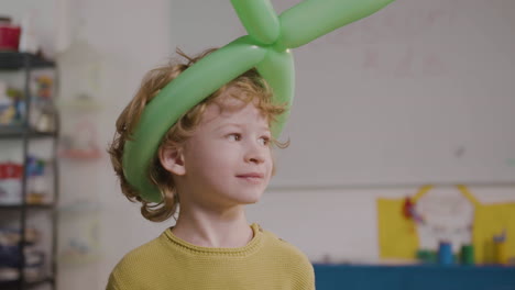 front view of a little boy with a long green balloon on his head in classroom in a montessori school