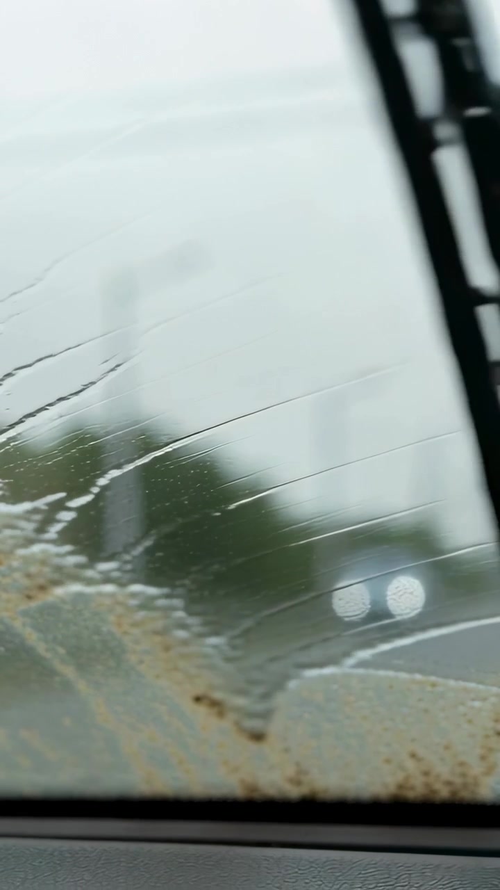 Video verticale: Attivazione dei tergicristalli, conducente che pulisce il vetro sporco di fango su strada bagnata, ripristinando la visibilità.