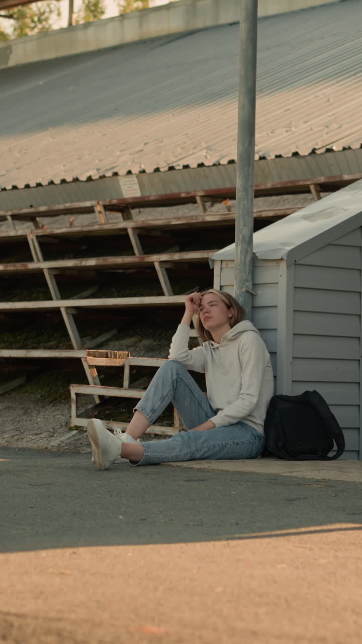 Young lady sits thoughtfully on stadium walkway, resting her head on her hand, leaning on a pole with her black bag beside her, she gazes upward, immersed in quiet reflection