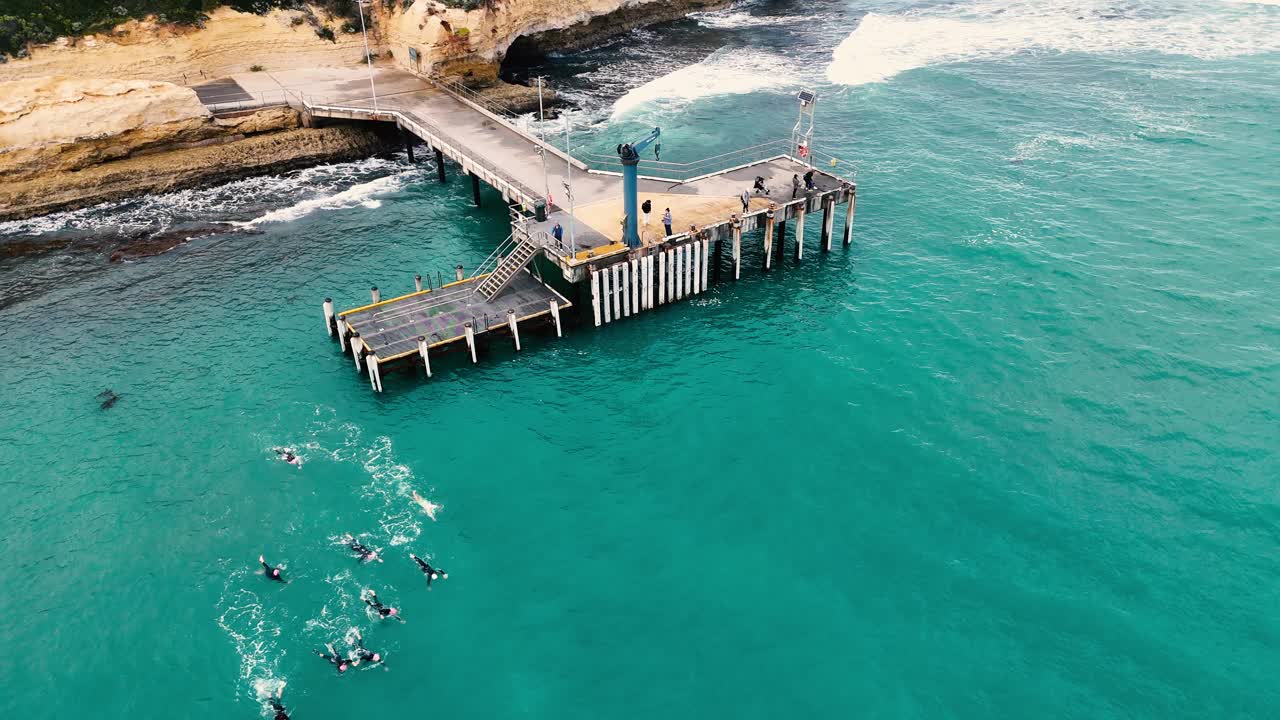 Drone captures swimmers training near Port Campbell jetty in turquoise waters under clear skies, showcasing teamwork and endurance