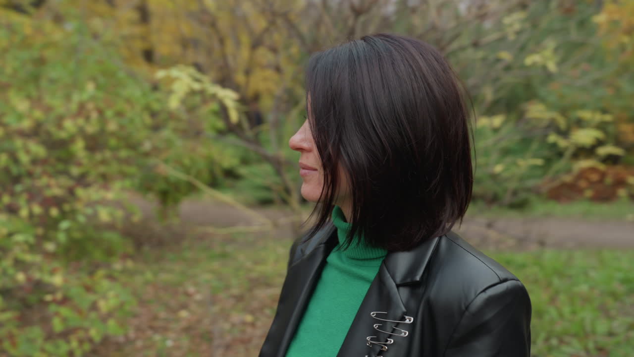 Donna con giacca di pelle in un parco autunnale, atmosfera rock contemplativa con dolcevita verde e caschetto, passeggiata spontanea e pose di profilo tra il fogliame dorato, luce diurna nuvolosa che crea un'atmosfera malinconica ma sicura di sé