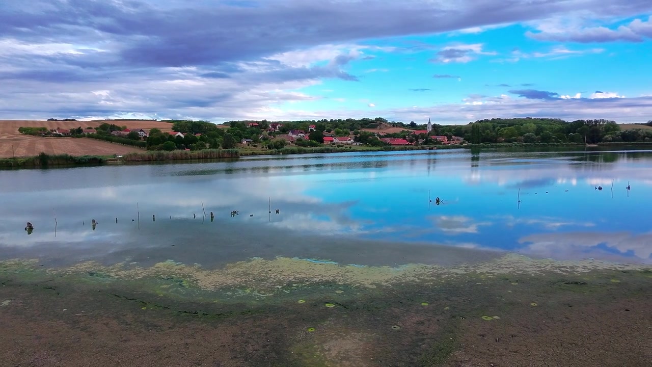 Una lenta e ampia panoramica della telecamera si muove attraverso un lago tranquillo che riflette un cielo azzurro brillante e nuvole, verso un pacifico villaggio rurale adagiato sulla collina ad Alsómocsolád, contea di Baranya, Ungheria