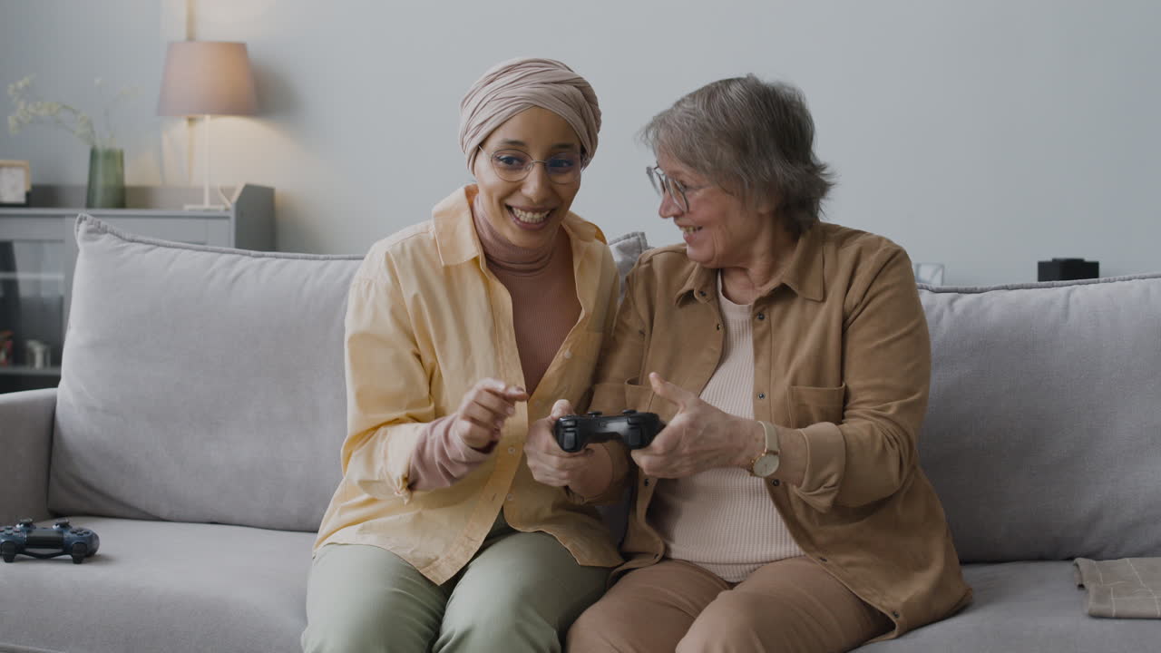 Middle Aged Arabic Woman Explaining To A Senior Lady How To Use The Game Controller While Sitting Together On Couch At Home