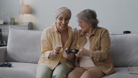 middle aged arabic woman explaining to a senior lady how to use the game controller while sitting together on couch at home