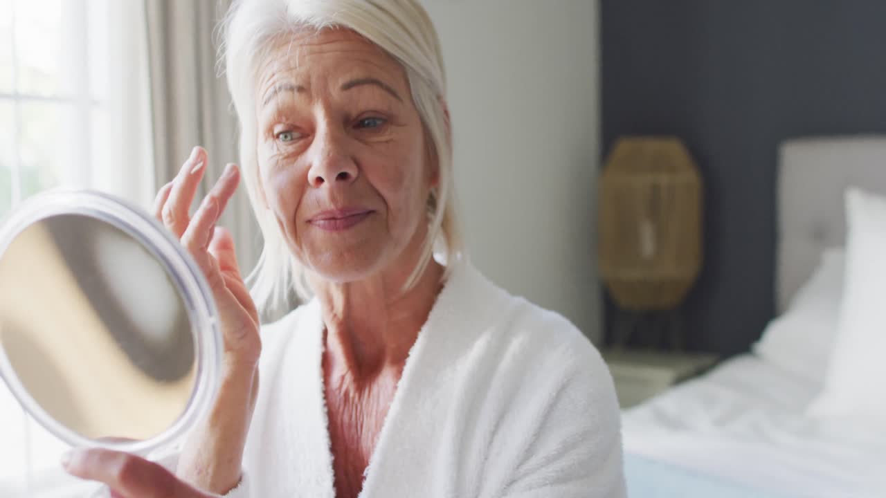 mujer caucásica mayor feliz sentada en la cama en el dormitorio, mirando al espejo, tocando su cara