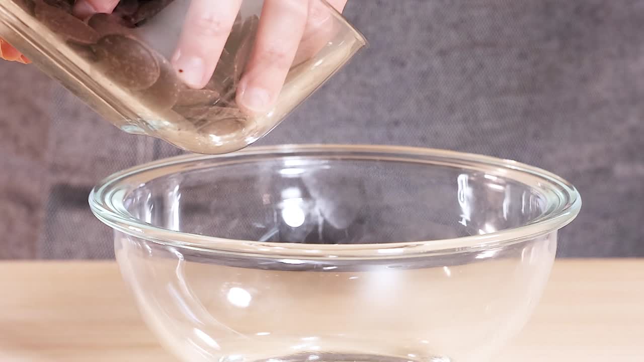 Close-up of chocolate discs being poured into a clear glass bowl on a wooden surface.