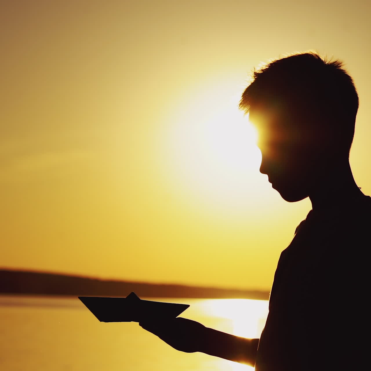 Silhouette di un ragazzo in piedi vicino al fiume che gioca con una barchetta di origami a un bellissimo tramonto. La barchetta di carta è nelle mani di un bambino piccolo e il sole splendente della sera