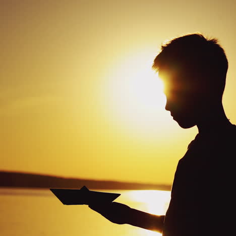 silhouette of a boy standing near the river and playing with origami ship at a beautiful sunset. paper boat is in the hands of a little child and bright evening sun