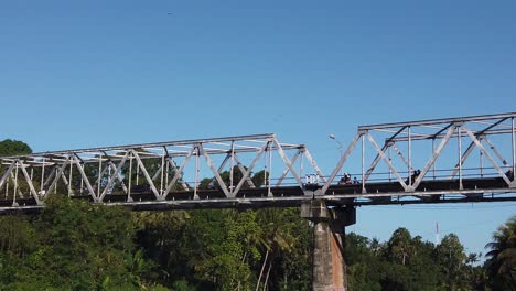 Motos-Y-Scooters-Conducen-Por-Encima-De-Un-Puente-De-Acero-Sobre-La-Selva-Tropical-Y-El-Horizonte-Azul-En-Klungkung,-Bali,-Indonesia,-Sudeste-De-Asia