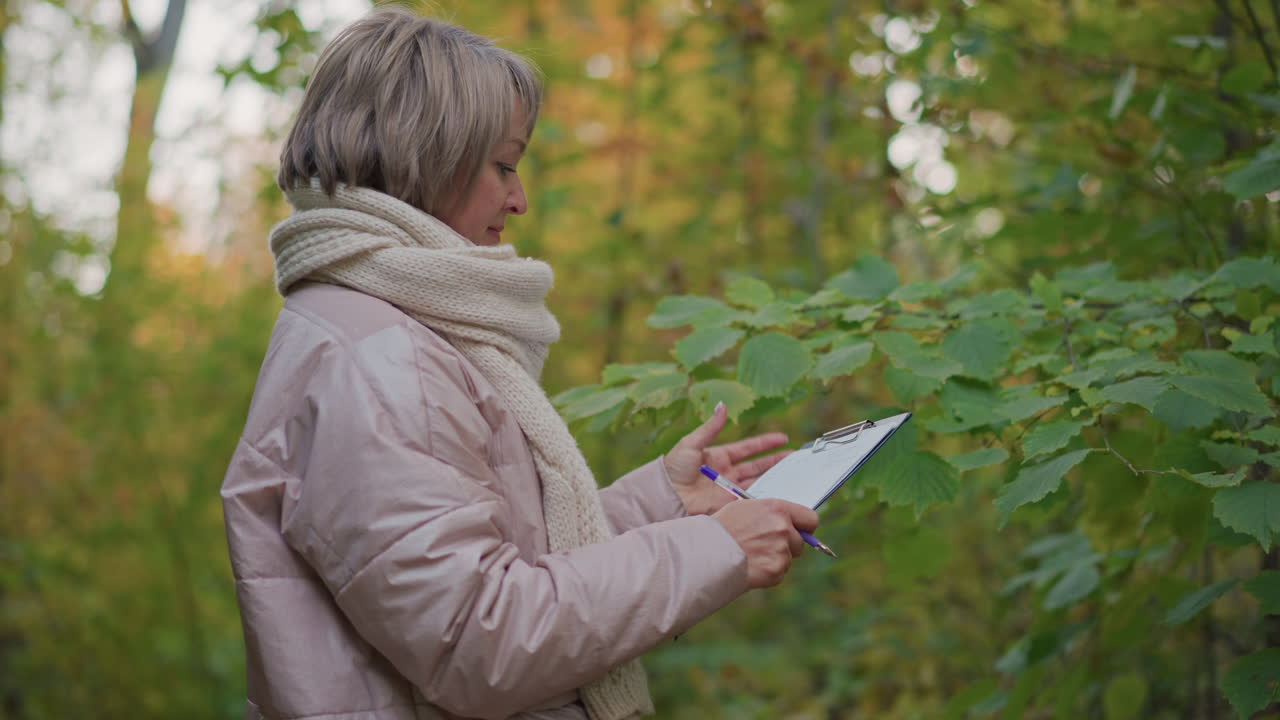 scientist wearing pink padded jacket and thick scarf holds clipboard and pen while carefully observing and examining green leaf in lush autumn forest during quiet daytime