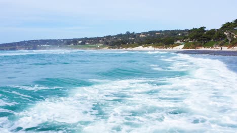 Aerial-Low-Flyover-of-Pacific-Ocean-waves-rolling-onshore-at-Carmel-by-the-Sea-Beach-with-Pebble-Beach-Golf-Course-in-background