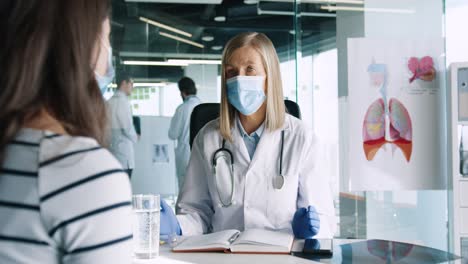 doctora caucásica con mascarilla médica sentada en el escritorio y explicando a una paciente el tratamiento del coronavirus en consulta médica