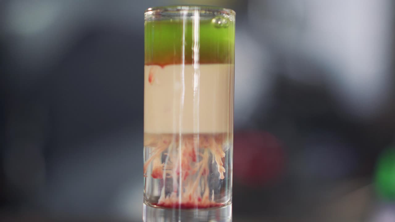 Barman adds red liquid into green and white cocktail into a glass on table. Red drops of alcohol falling in the cocktail