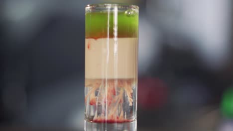 barman adds red liquid into green and white cocktail into a glass on table. red drops of alcohol falling in the cocktail