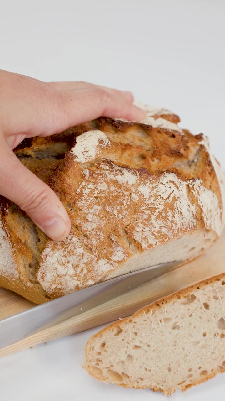 Caucasian hands use a serrated knife to slice crusty sourdough bread on a white background.