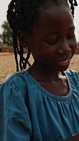 african girl wearing a blue dress examines a plastic water bottle in a rural village setting, her braided hair and small earrings visible as she looks down