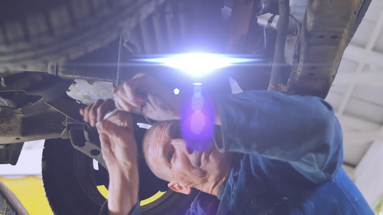 senior male mechanic lying underneath vehicle in auto garage, showing animated diagnostic icons