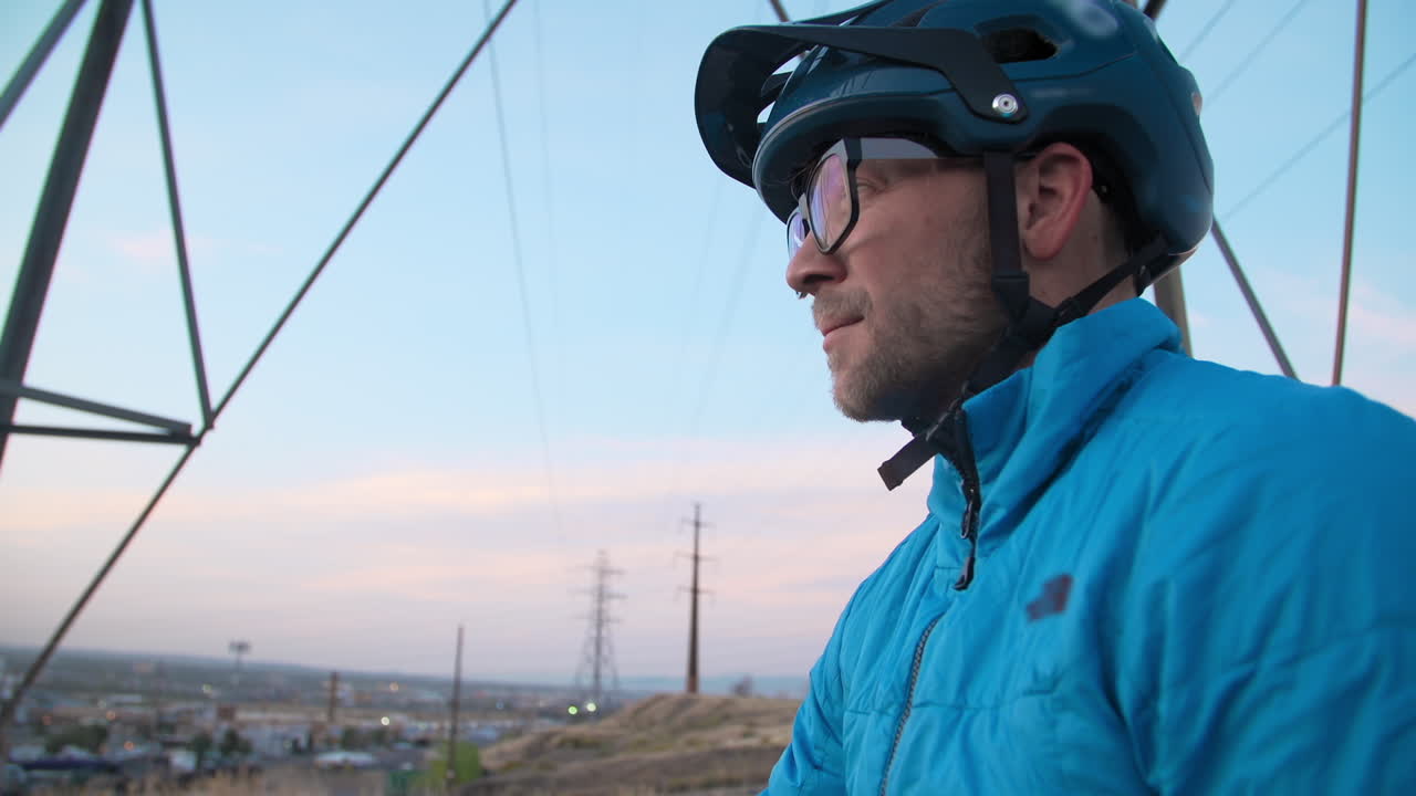 toma de carro medio de un ciclista de montaña masculino, en la cima de una colina observando el horizonte de una ciudad debajo de una torre de línea eléctrica mientras el sol bajo crea un cielo dramático