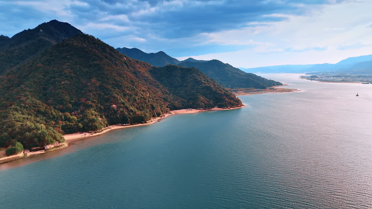 Narrow line of the sandy beach located at the foot of the mountain range covered with thick woods. Aerial perspective on the stunning calm waterscape reflecting bright sunlight.