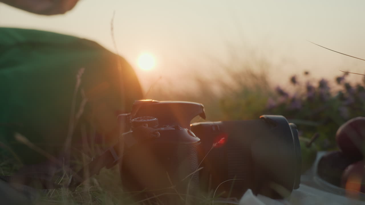 Close up of hand reaching for wine glass resting on grassy ground during golden hour sunset picnic, with visible fruit, flowers, and warm natural light