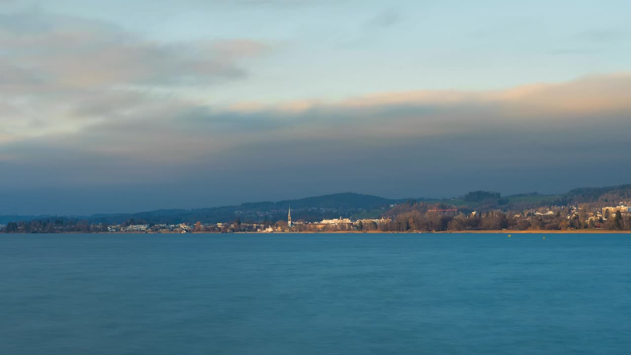 Time lapse of a cloudy sunset on a lake. At the back you can see a small town with a church in the center. It is cold on the lake called Pfäffikersee. The sky is partly blue. Sun illuminate the Town.