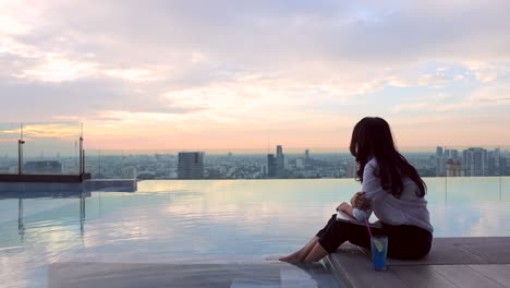 asian businesswoman sitting on the ledge of the pool with her feet in the water. woman is working, typing on her laptop and enjoy view skyscraper in city with beautiful sunset or sunrise sky.