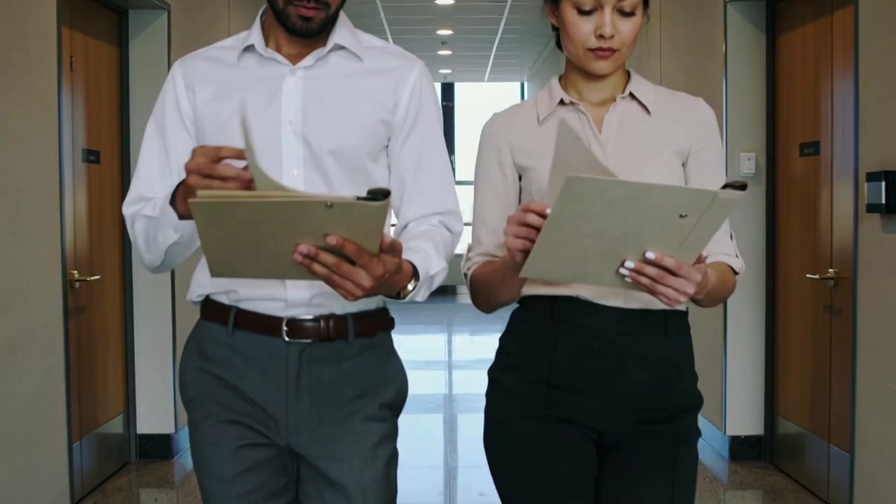 Hombre y mujer caminando con ropa de negocios revisando archivos beige en un pasillo, la cámara comenzando