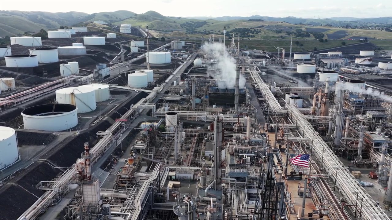 Phillips 66 San Francisco Refinery in Rodeo, California, with large American flag waving over industrial pipes and white storage tanks under overcast sky. Aerial drone shot.