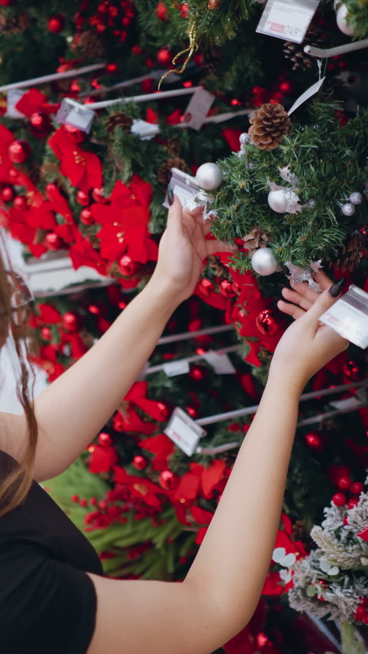vista aérea de una mujer joven de cabello largo y marrón con camisa negra admirando las coronas de navidad decoradas con adornos de plata, piñas y cintas rojas en el pasillo de la tienda de vacaciones