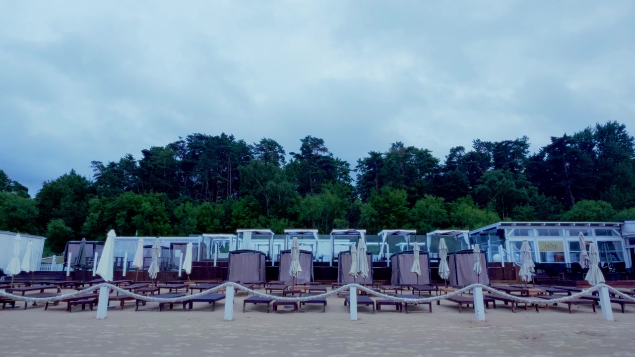 Ampia inquadratura statica che mostra un lounge bar vuoto sul mare con lettini prendisole, tende cabana bianche e una recinzione in corda lungo la spiaggia sabbiosa del Baltico nella località di Jurmala, Lettonia