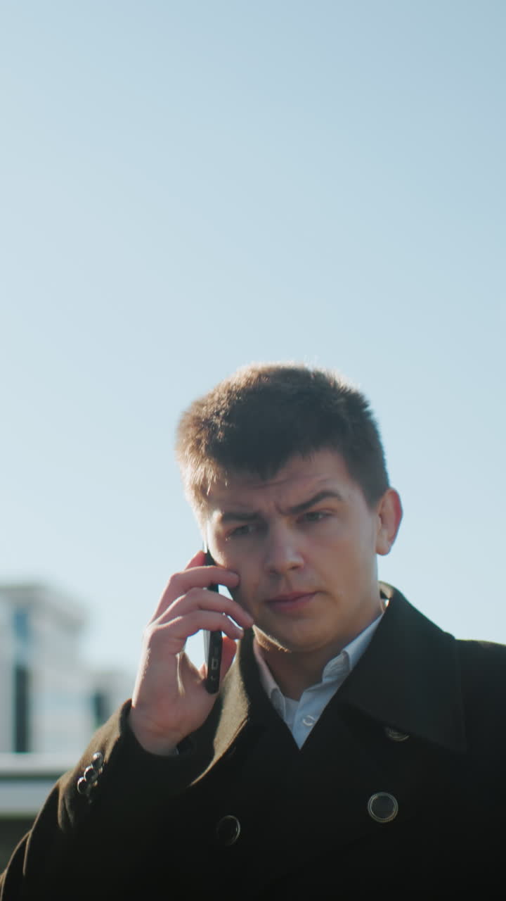 금속 버튼으로 된 검은 코트를 입은 사업가, 은 파란 하늘 아래 밖에서 전화 통화, 현대 도시 환경에서 주차 된 자동차 근처에서 는 동안 불쾌하고 상하는 것처럼 보입니다.