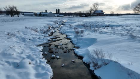 snowy landscape with a meandering stream near a farm under a cloudy sky