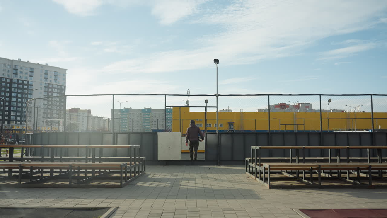Back view of person walking away from urban sports arena, carrying soccer ball, with vibrant yellow storage containers and city buildings in the background