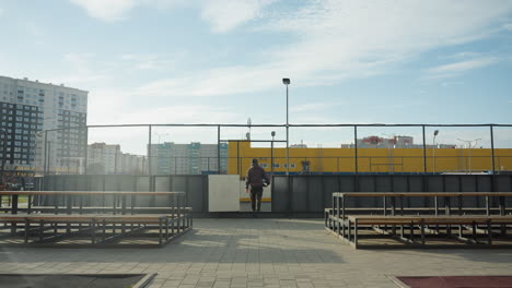back view of person walking away from urban sports arena, carrying soccer ball, with vibrant yellow storage containers and city buildings in the background