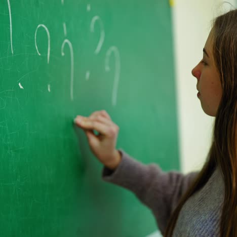 student girl at school. side view of a schoolgirl standing near the blackboard and writing with a white chalk. teenager writes question marks on a board.