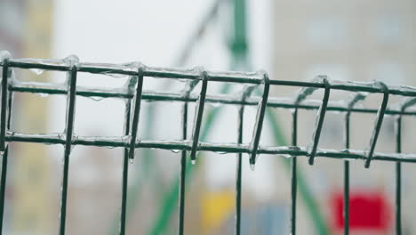 close-up of a green chain-link fence with ice formations dripping from the bars, set against a blurry snowy park background, capturing a serene winter scene