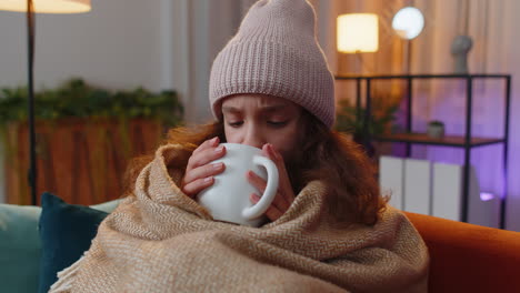 Sick-preteen-girl-wear-hat-wrapped-in-plaid-sit-alone-shivering-from-cold-on-sofa-drinking-hot-tea