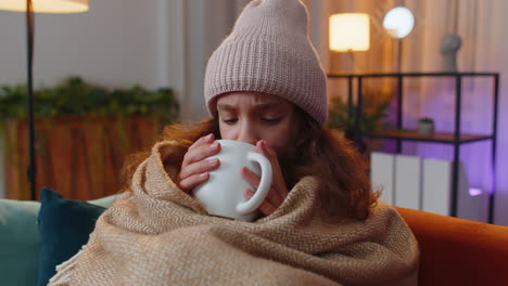 sick preteen girl wear hat wrapped in plaid sit alone shivering from cold on sofa drinking hot tea