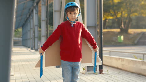 cute little boy in helmet and red sweater with cardboard airplane wings looking at camera and pretending to fly