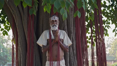 White beaded sadhu meditating, forehead marked with sacred markings, sitting beneath tree adorned with crimson ritual threads in traditional spiritual setting